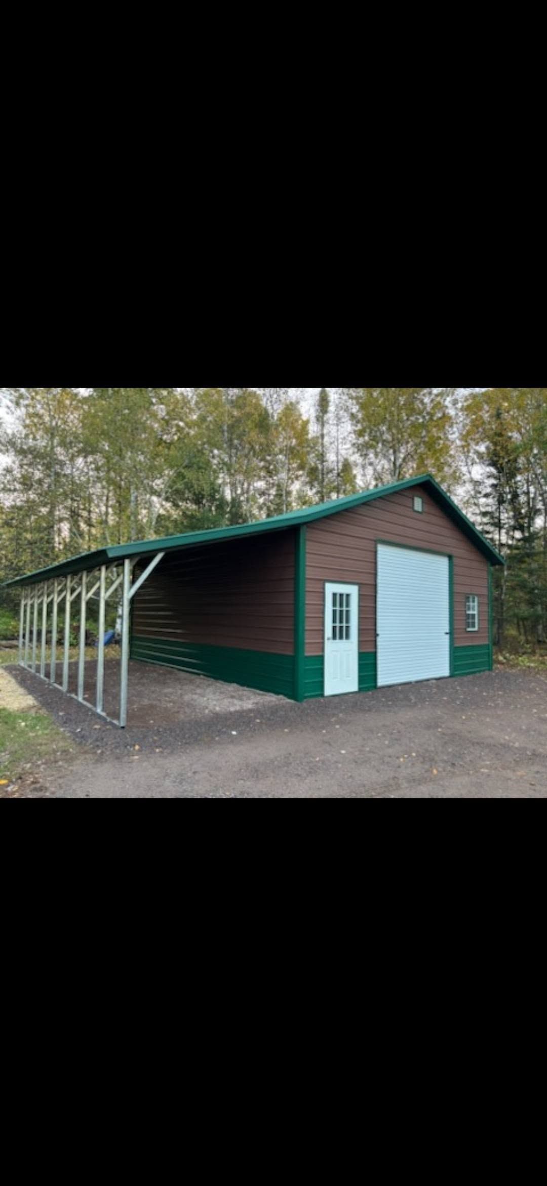 Green metal barn with a white roll-up door and covered side overhang in a wooded area