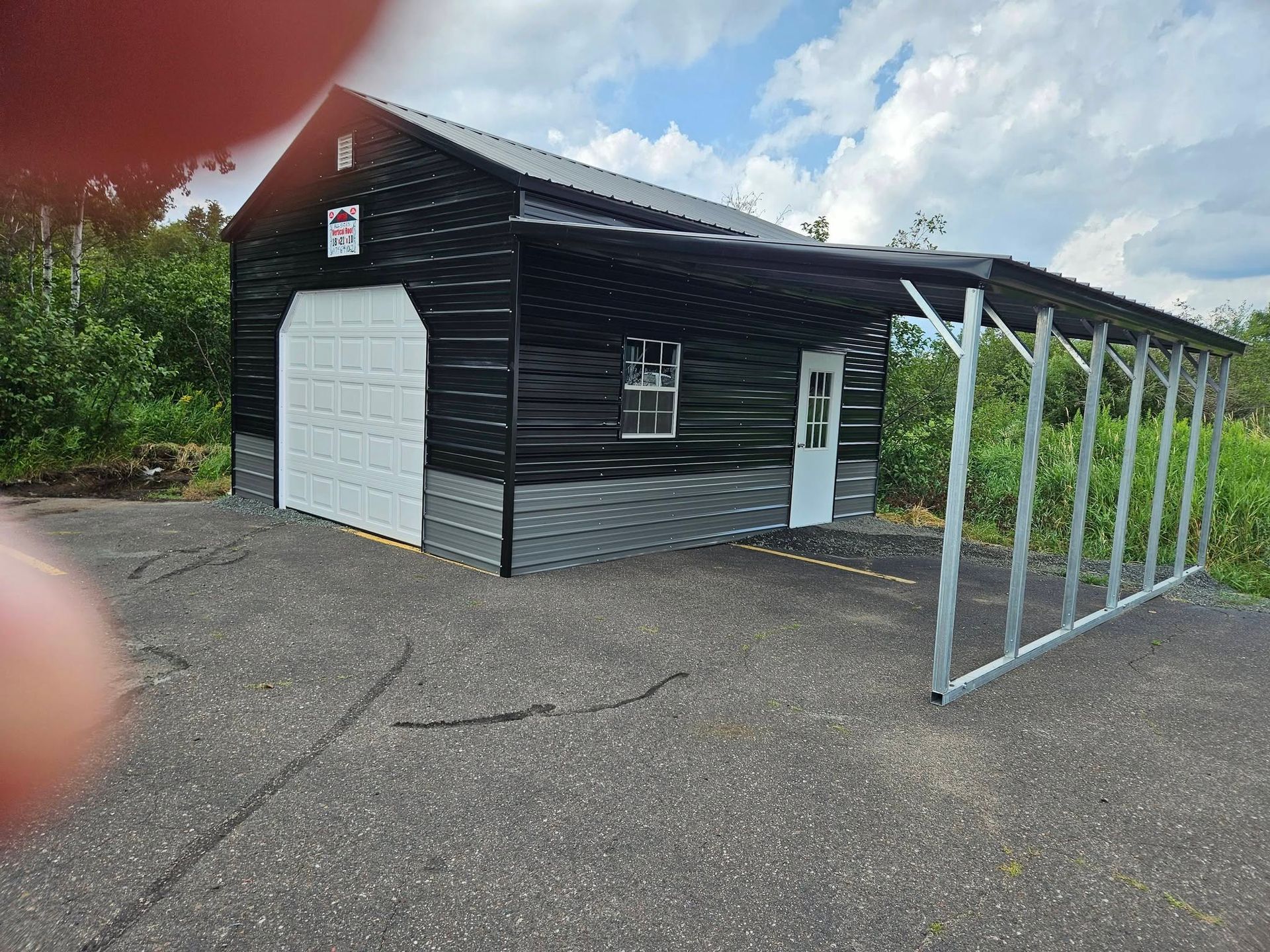 Black shed with white garage door and metal carport on a paved lot under a cloudy sky