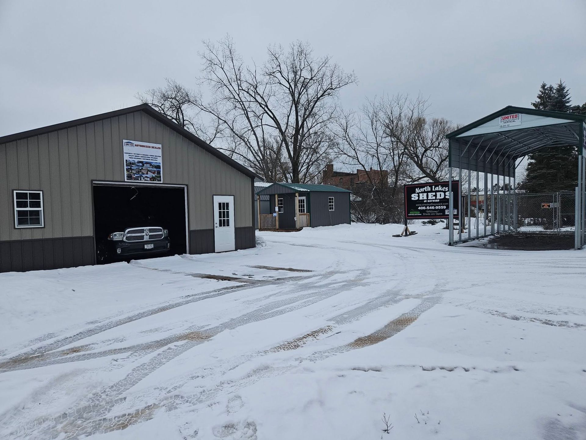 Snowy driveway beside a metal garage and open shed under bare trees.