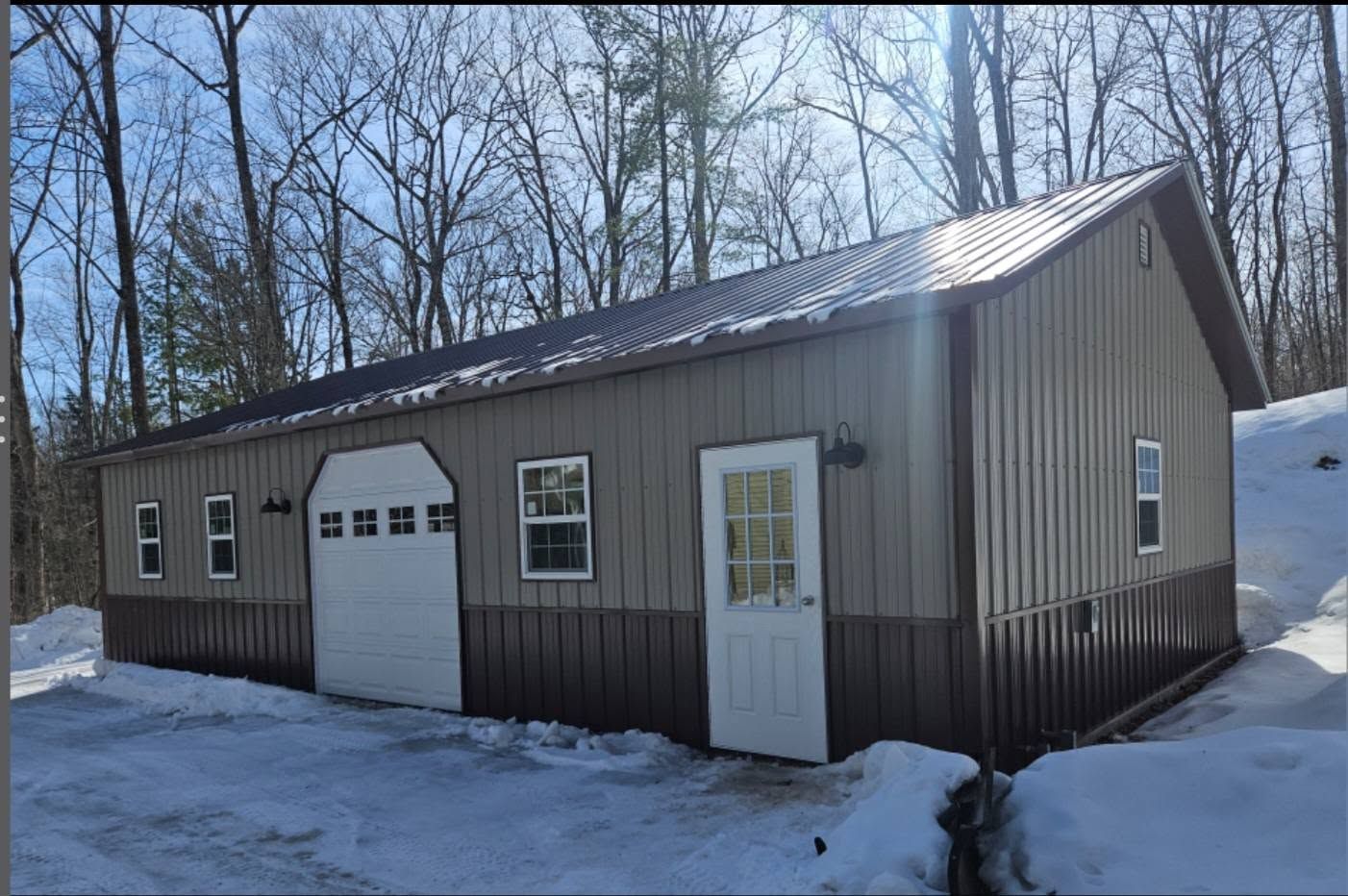 Gray metal garage with white doors and windows, set beside snowy ground and leafless trees