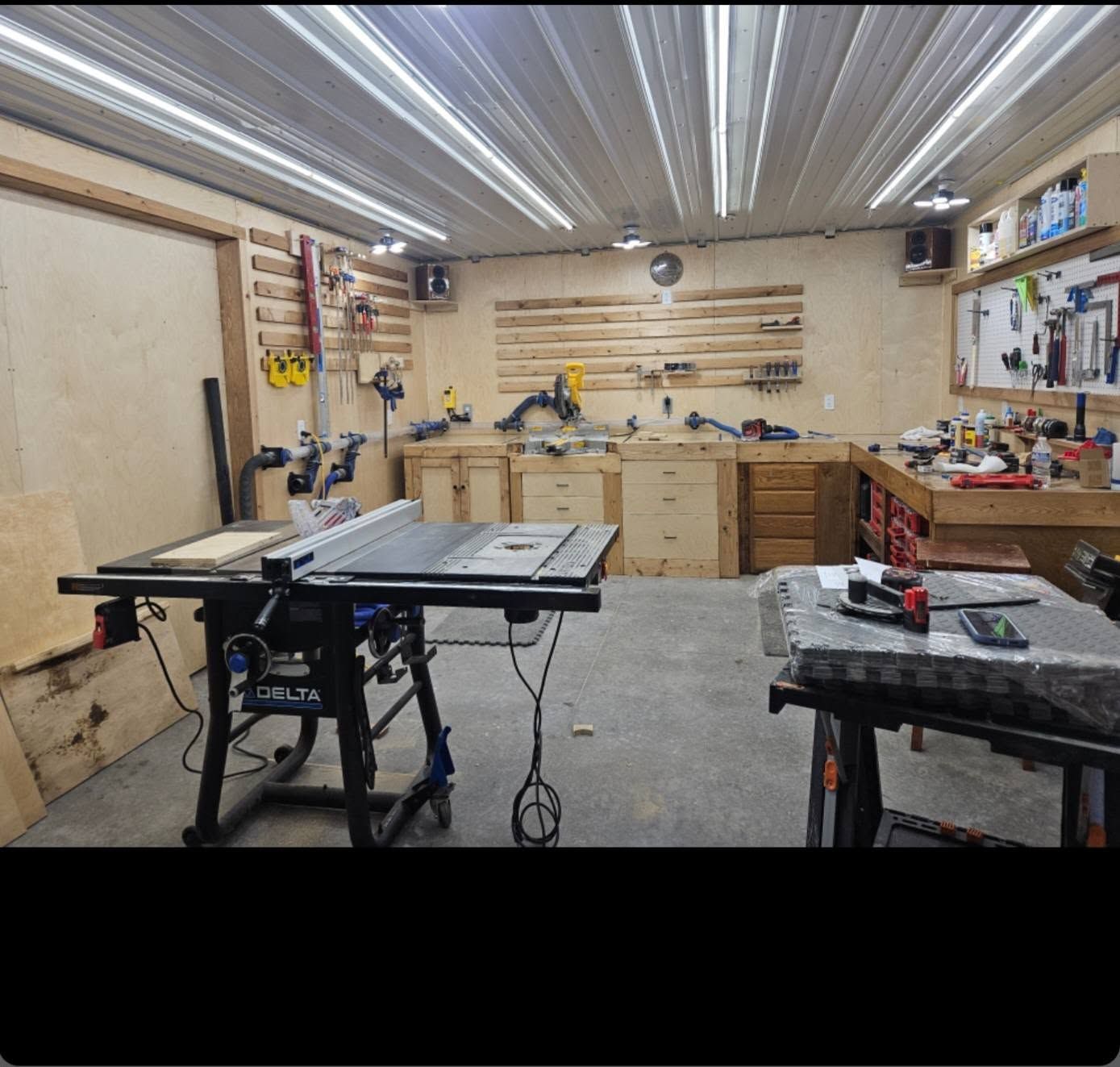 Bright woodworking workshop with tools, workbenches, shelves, and a table saw under fluorescent lights