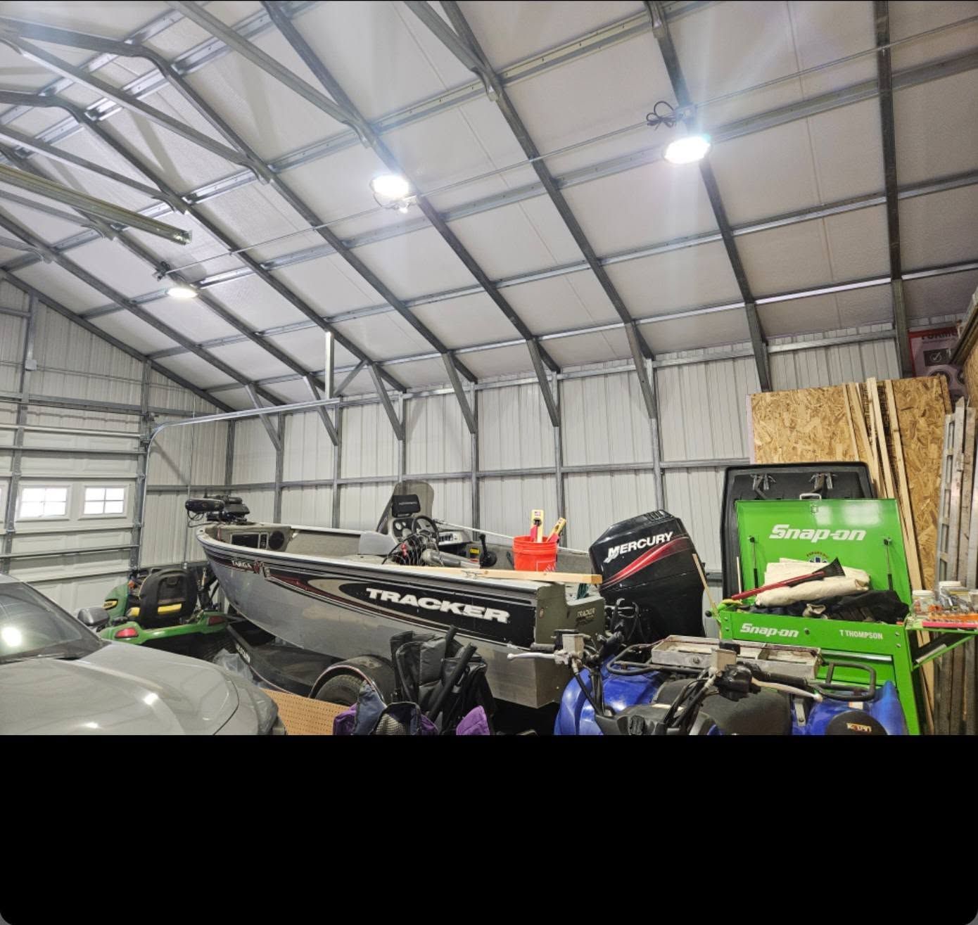Boathouse interior with bass boats, ATVs, a green machine, and stacked wood under a metal roof