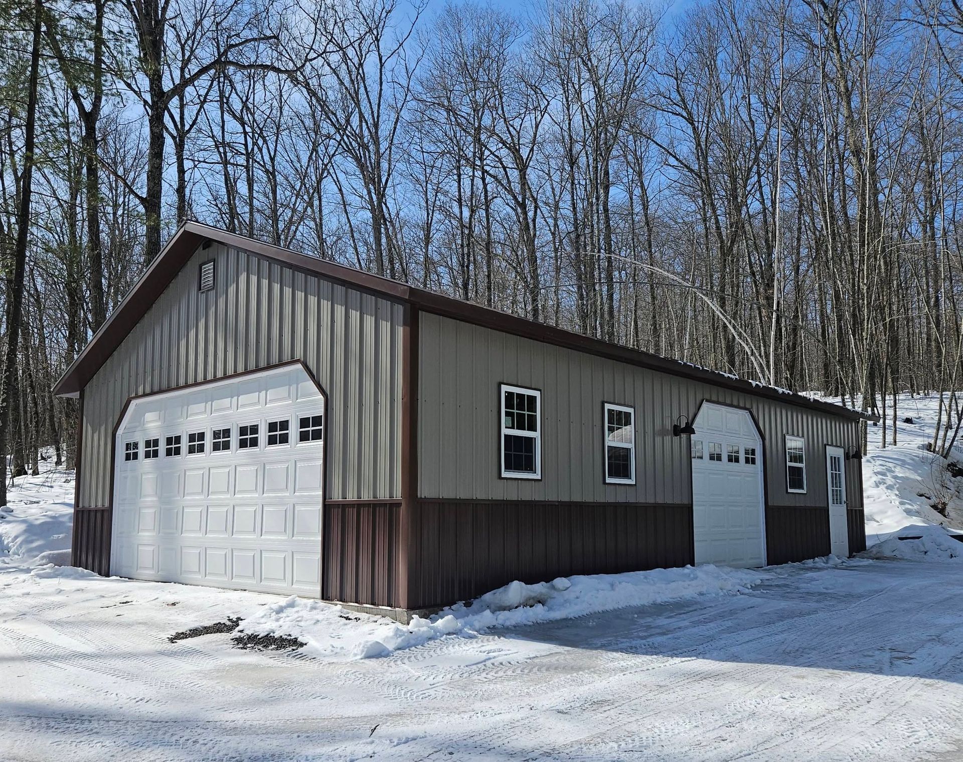 Brown metal garage building with white doors on a snowy wooded lot