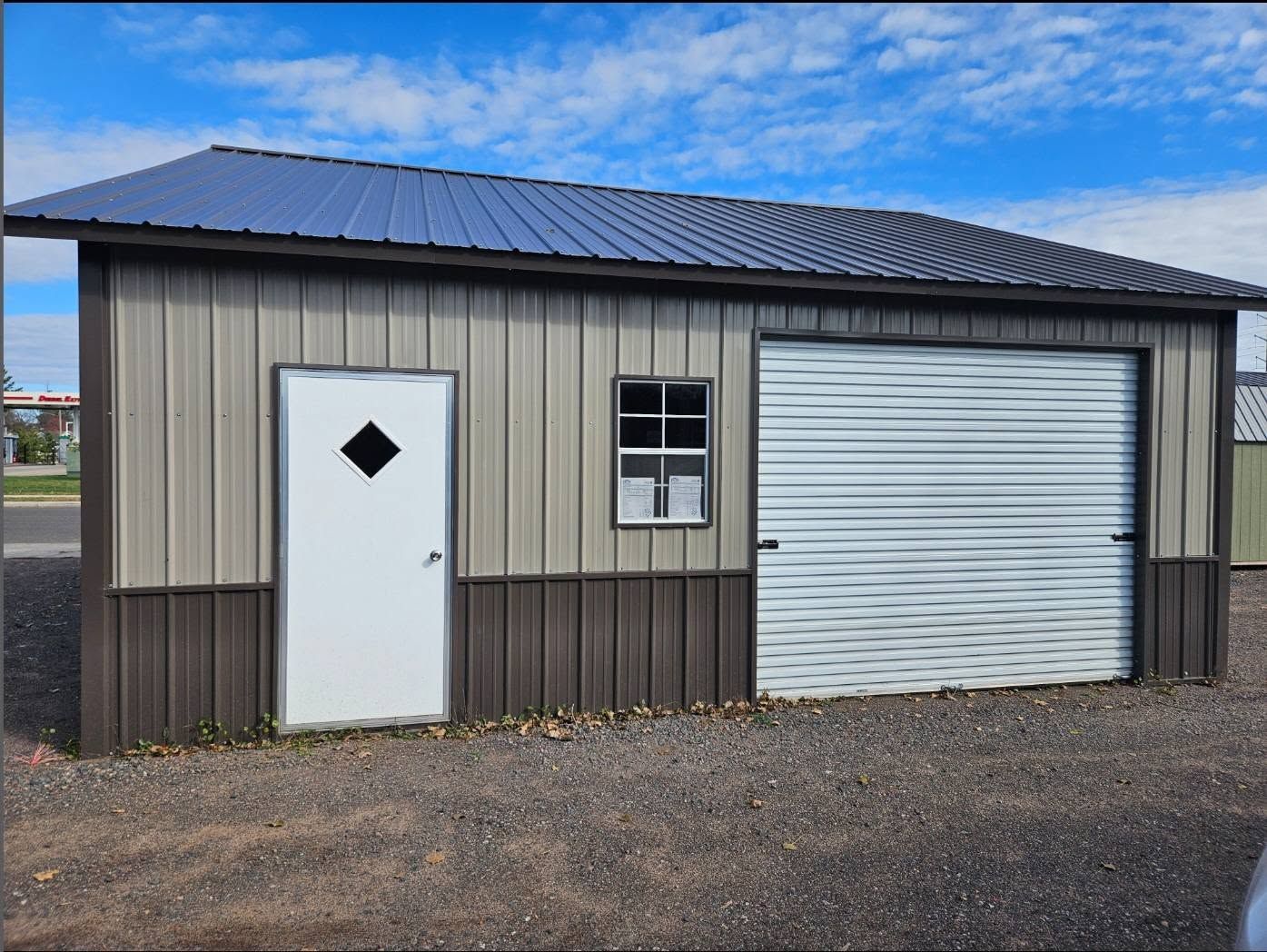 Small beige utility shed with a white door, window, and roll-up garage door on gravel.
