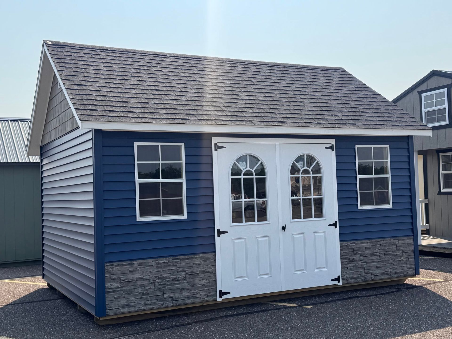 Small blue shed with white double doors and gray shingled roof in a yard.