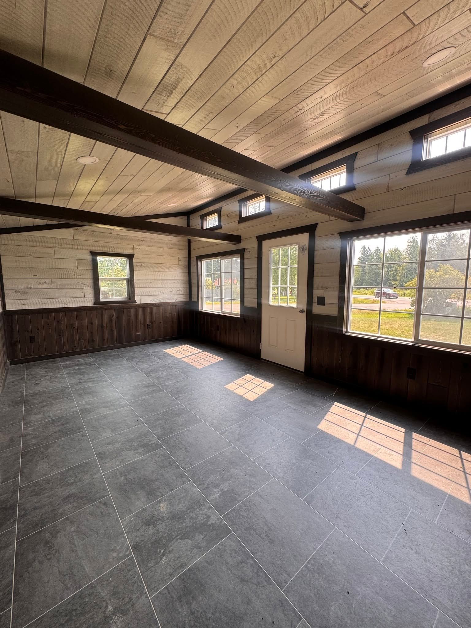 Empty sunlit room with wood-paneled walls, exposed ceiling beams, and large windows.