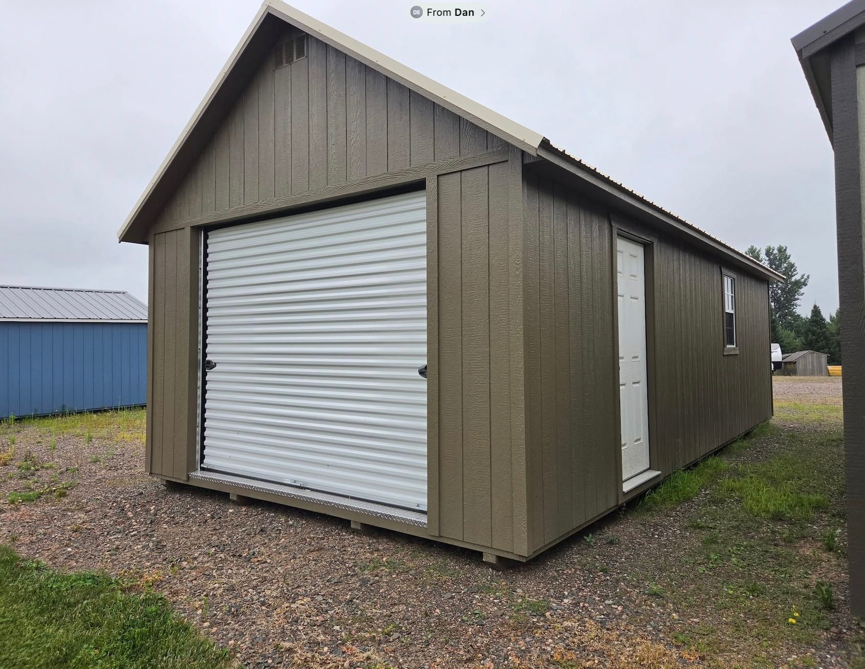 Small tan storage shed with a white roll-up door on a gravel lot under a cloudy sky