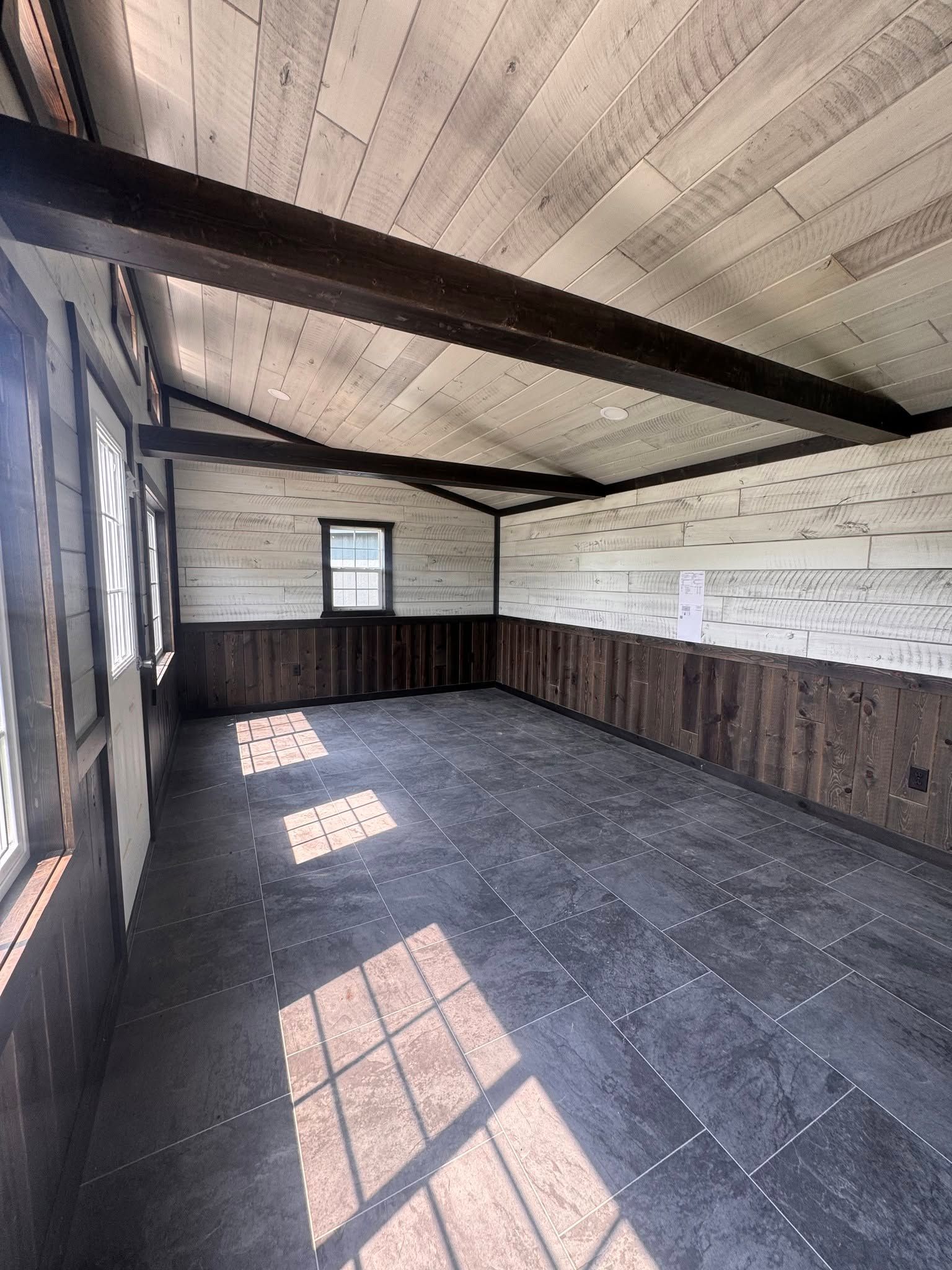 Empty rustic room with dark wood walls, slanted ceiling, and sunlight casting window shadows on the floor