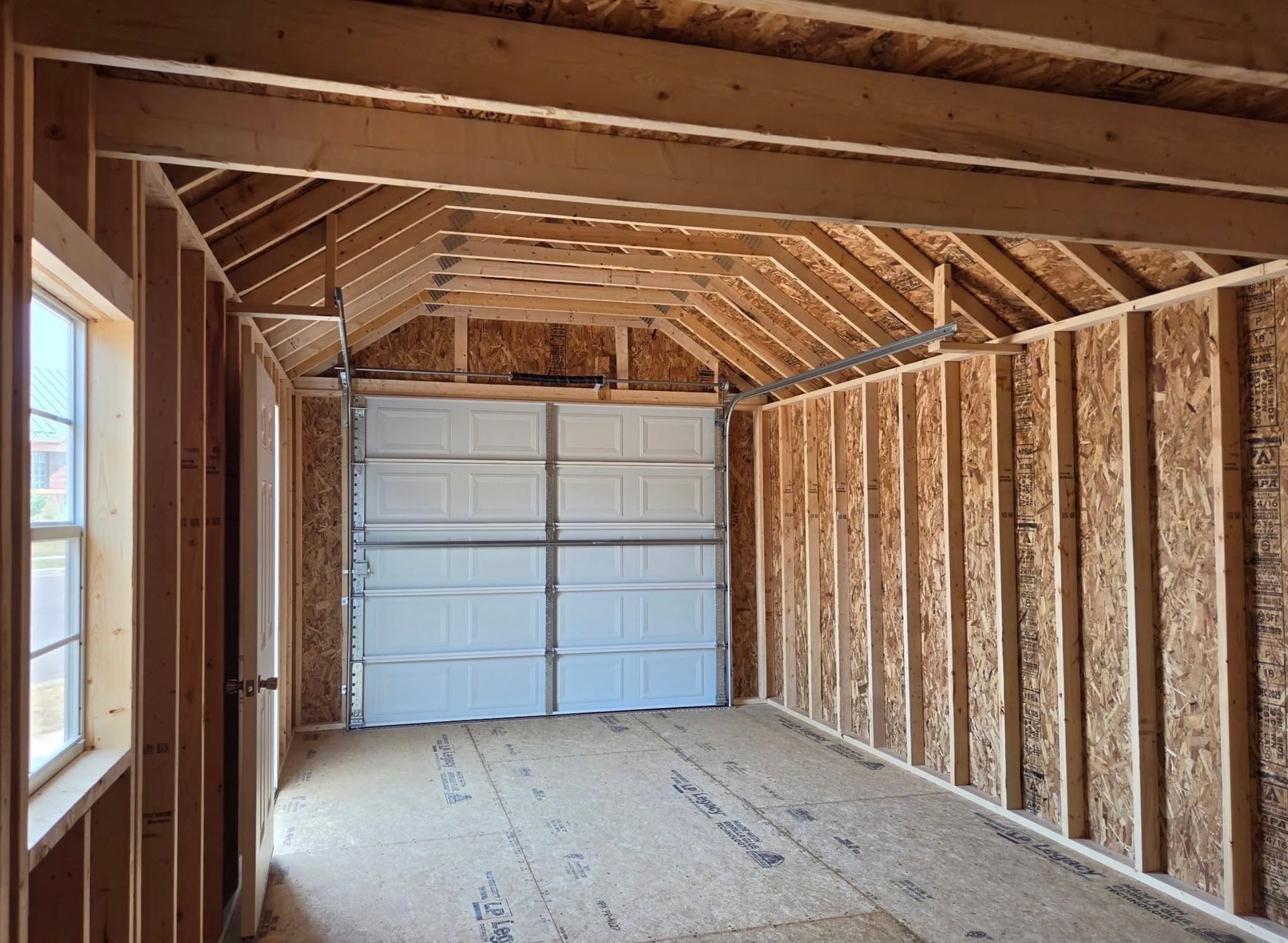 Empty framed garage interior with exposed rafters and a white roll-up door at the far end