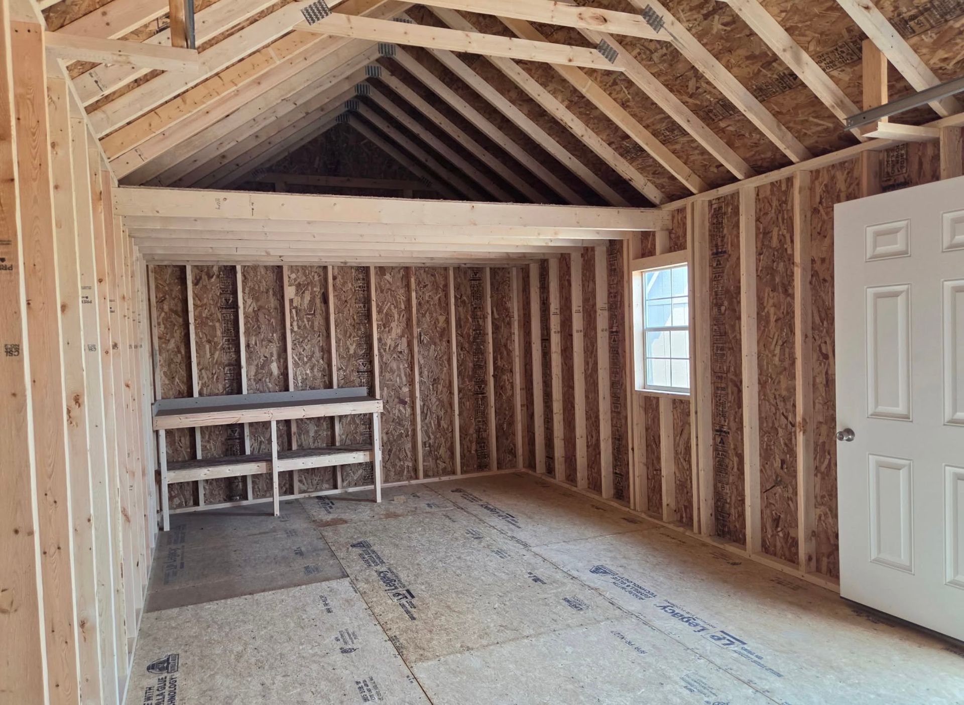 Unfinished attic room with exposed wooden framing, white door, small window, and shelf against the far wall