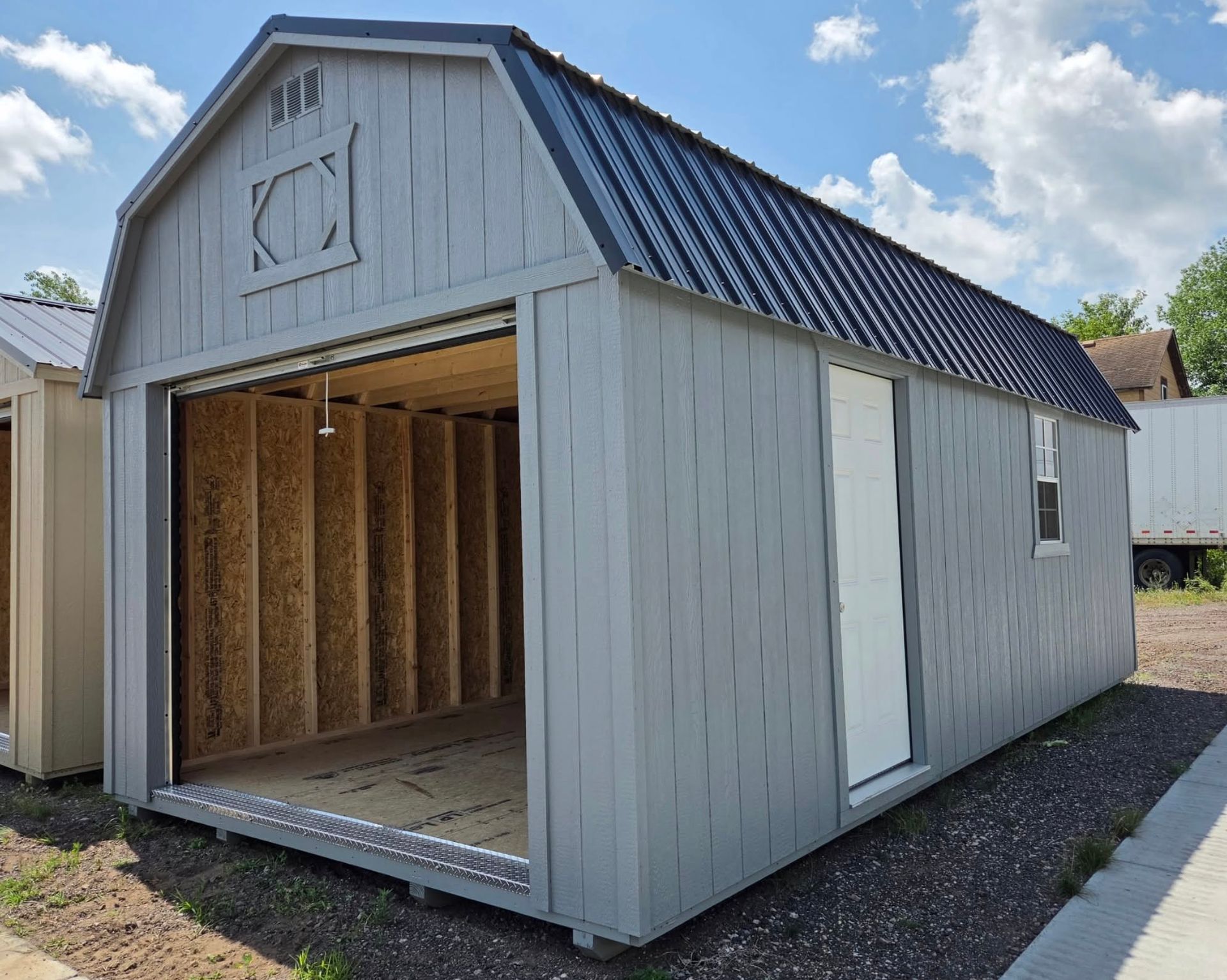 Small gray shed with open front, dark metal roof, and unfinished wood interior on a gravel lot