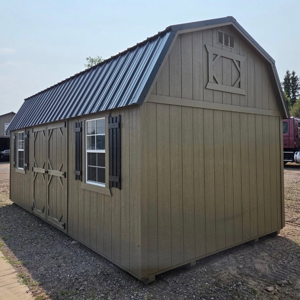 Small tan shed with a dark metal roof and shuttered windows, sitting on gravel outdoors.