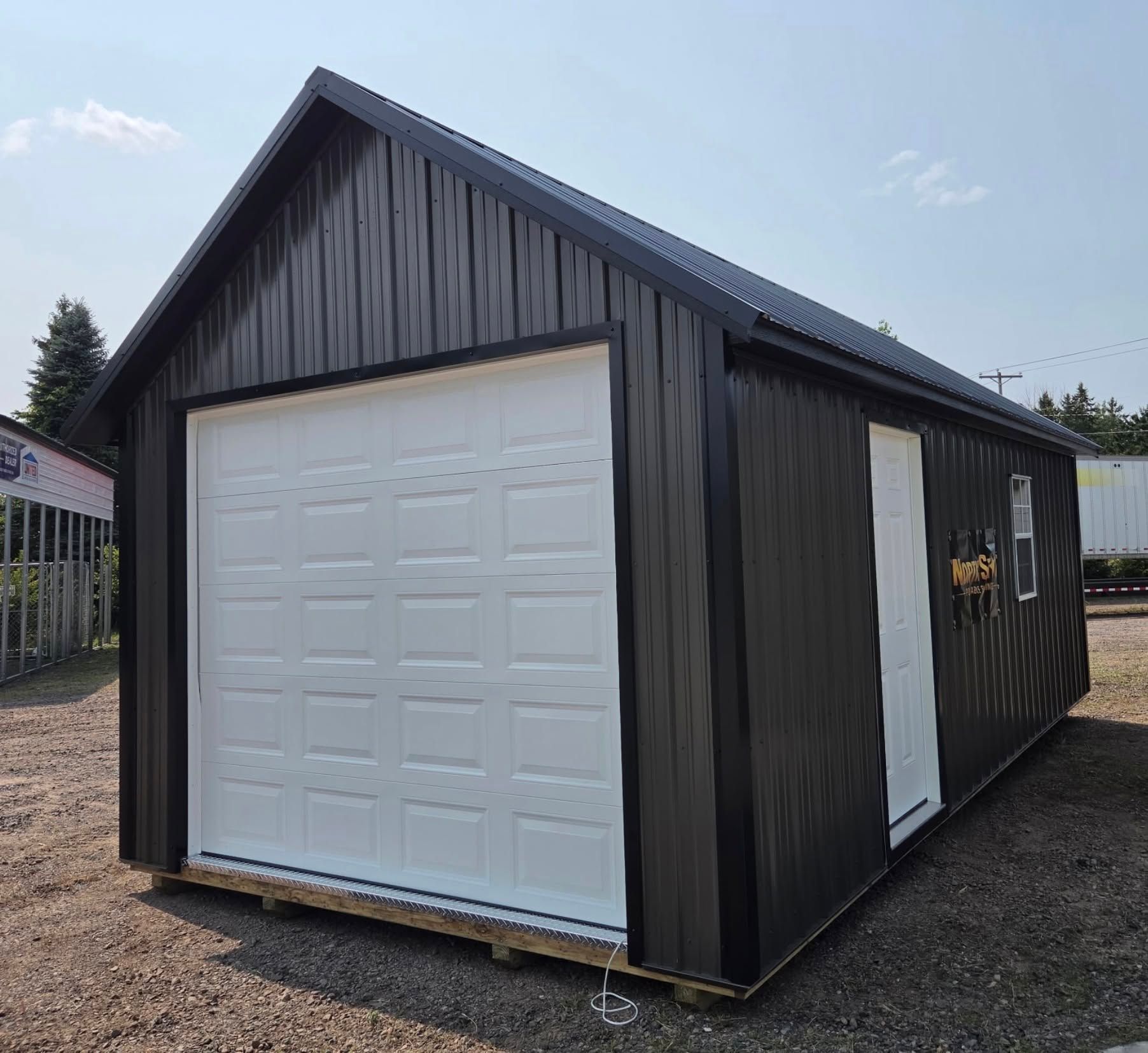 Black metal garage shed with a white roll-up door on a gravel lot under a partly cloudy sky