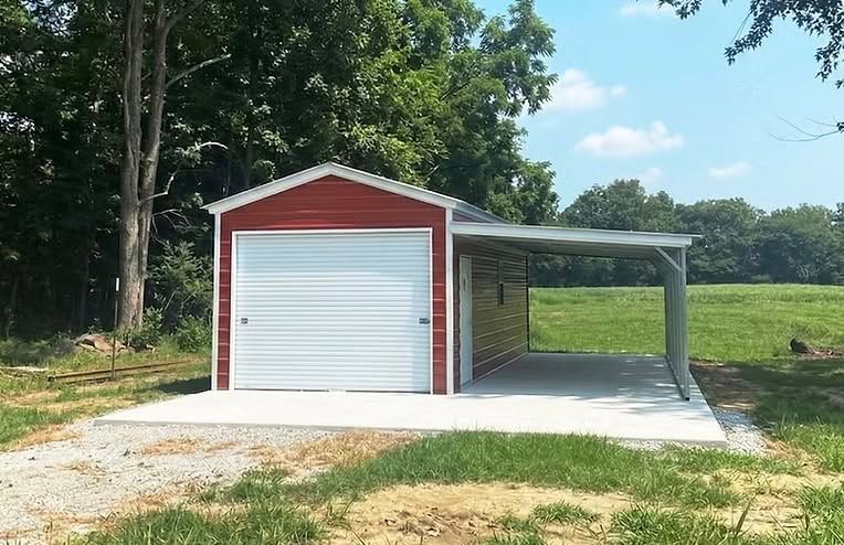 Red metal garage with white roll-up door and side carport on a gravel lot, surrounded by trees.