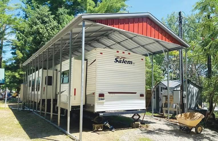 White Salem travel trailer under a metal carport in a wooded campsite, with a wheelbarrow nearby.