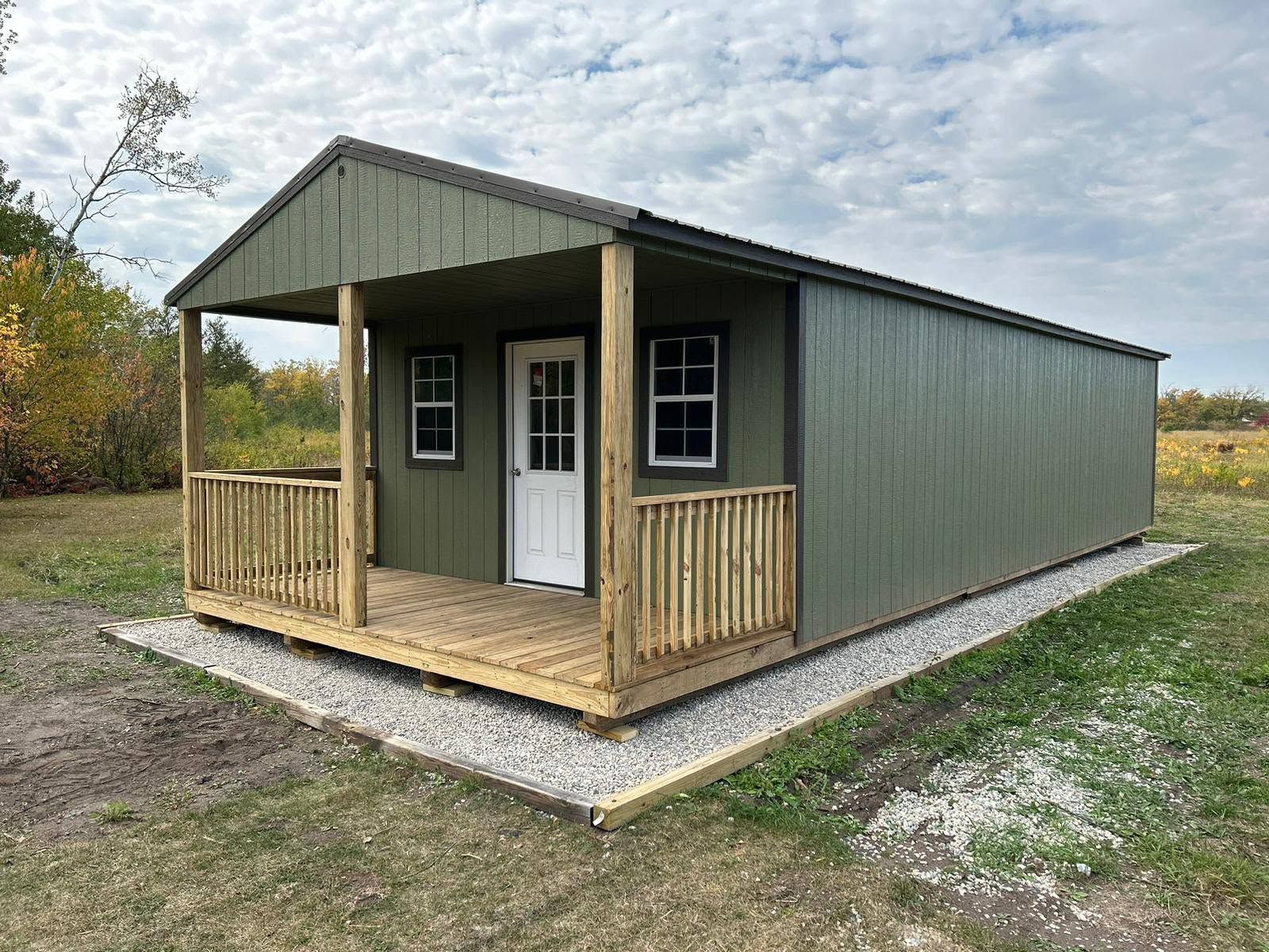 Small green cabin with a porch on a gravel pad in a rural field.