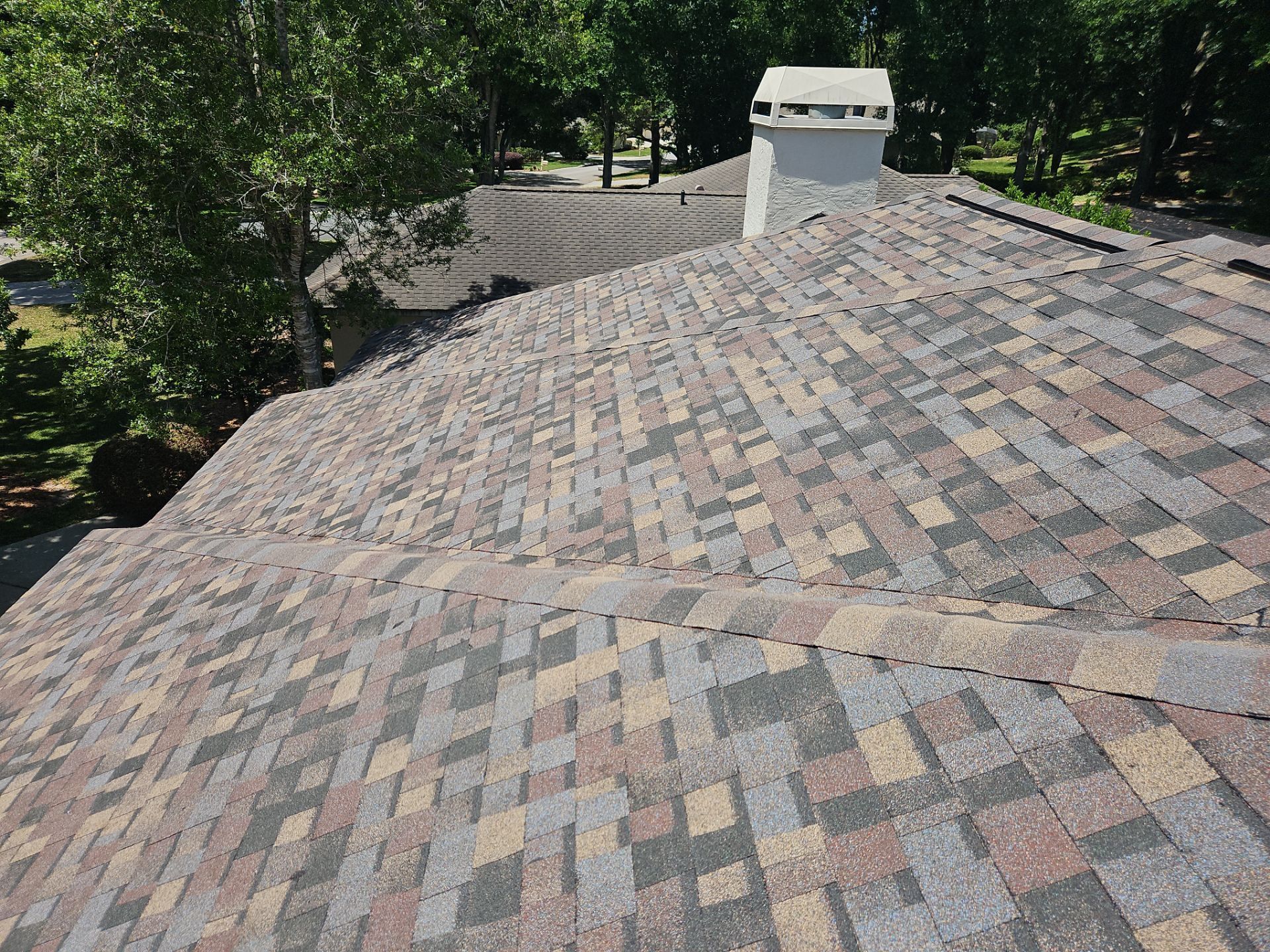 A multi-colored shingle roof with a chimney and trees in the background.