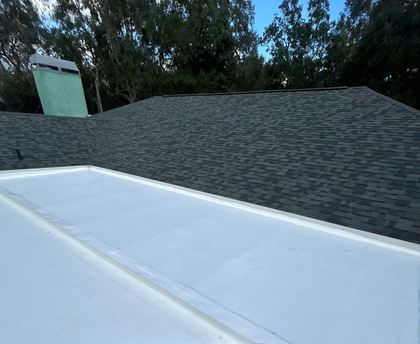 A flat roof with black shingles and a flat, white section, with a green chimney in the background.