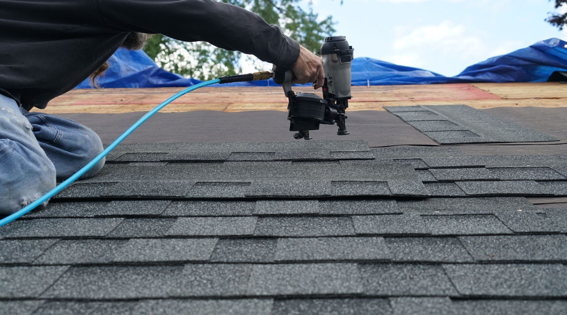 Person using a nail gun to install asphalt shingles on a roof, blue air hose attached.