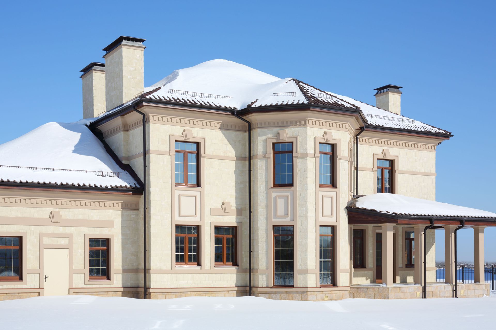 Two-story beige brick house covered in snow against a bright blue sky.
