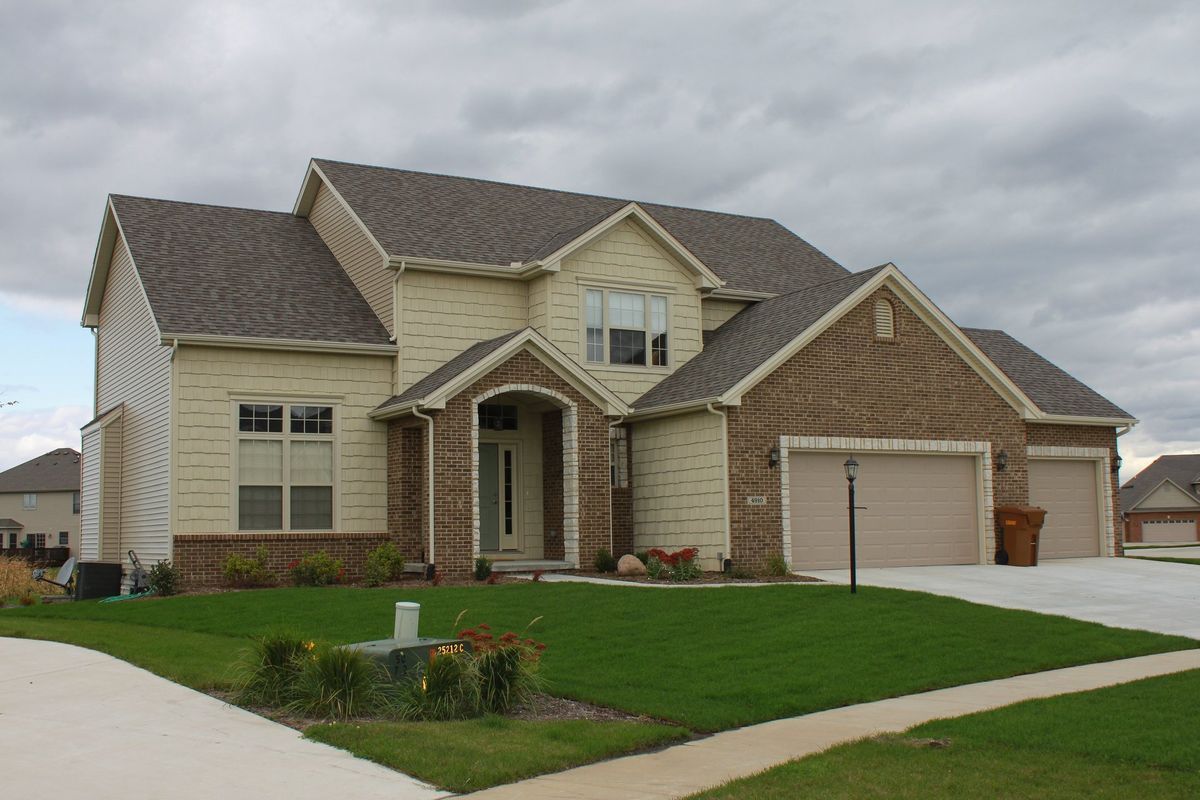 Two-story house with tan siding, brick accents, three-car garage, and green lawn under a cloudy sky.