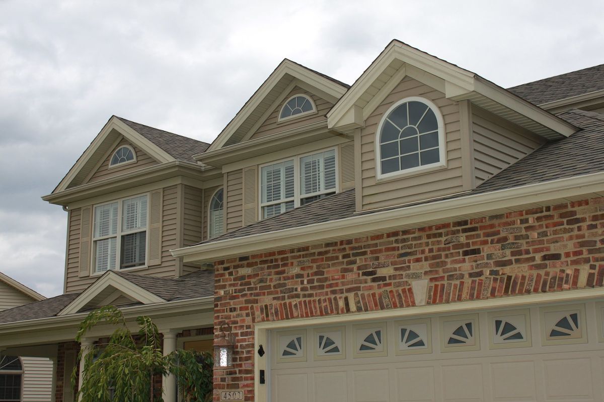 Two-story house with brick and tan siding, garage door, and multiple windows under a cloudy sky.