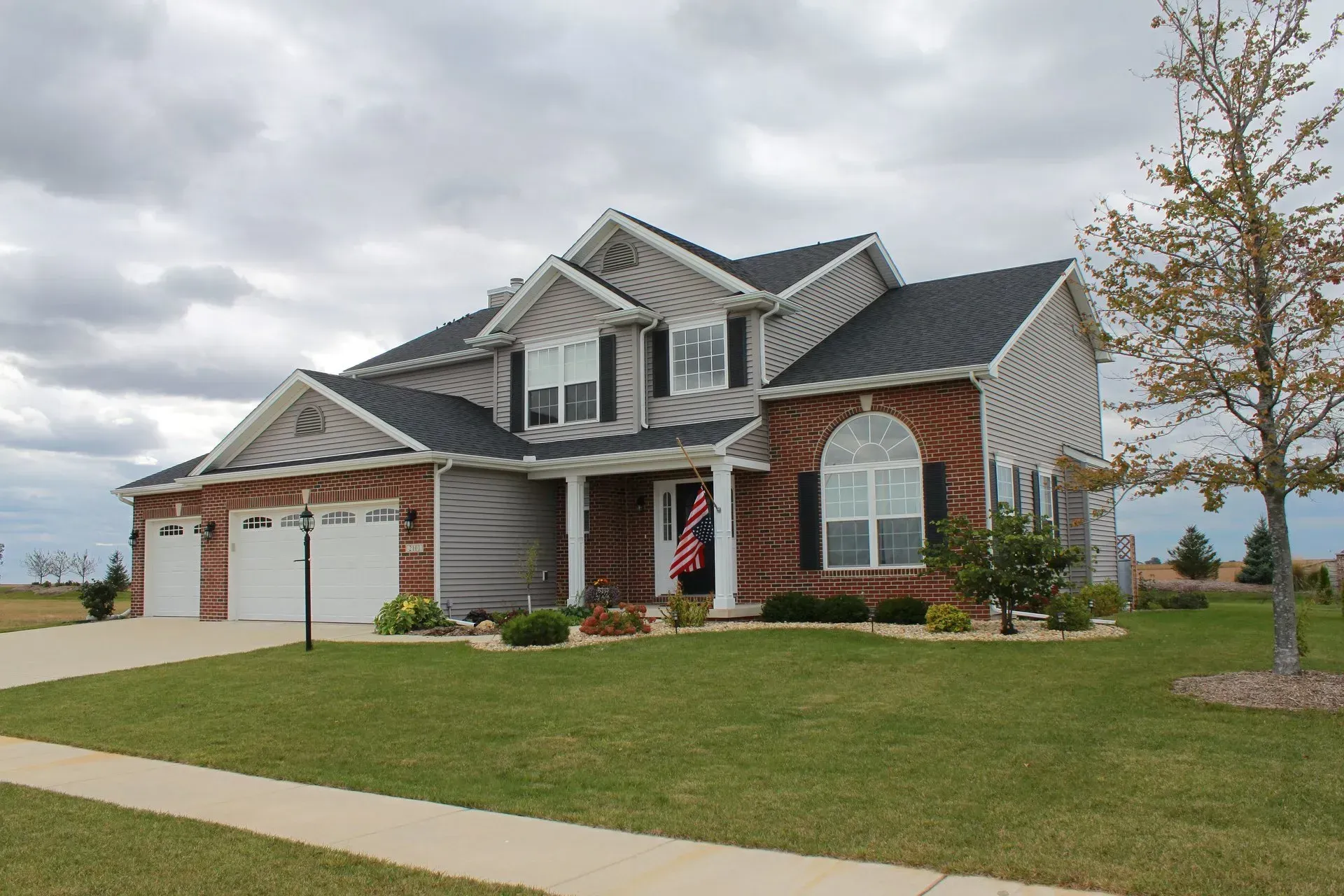 Two-story brick house with a dark roof and a well-manicured lawn under a cloudy sky.