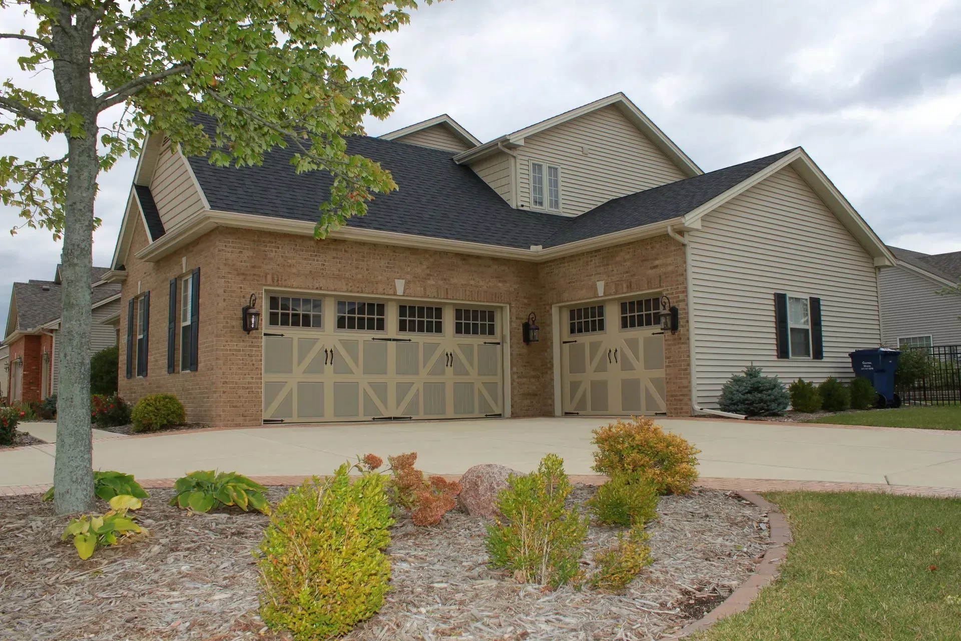Two-story brick house with beige garage doors, a black roof, and manicured landscaping.