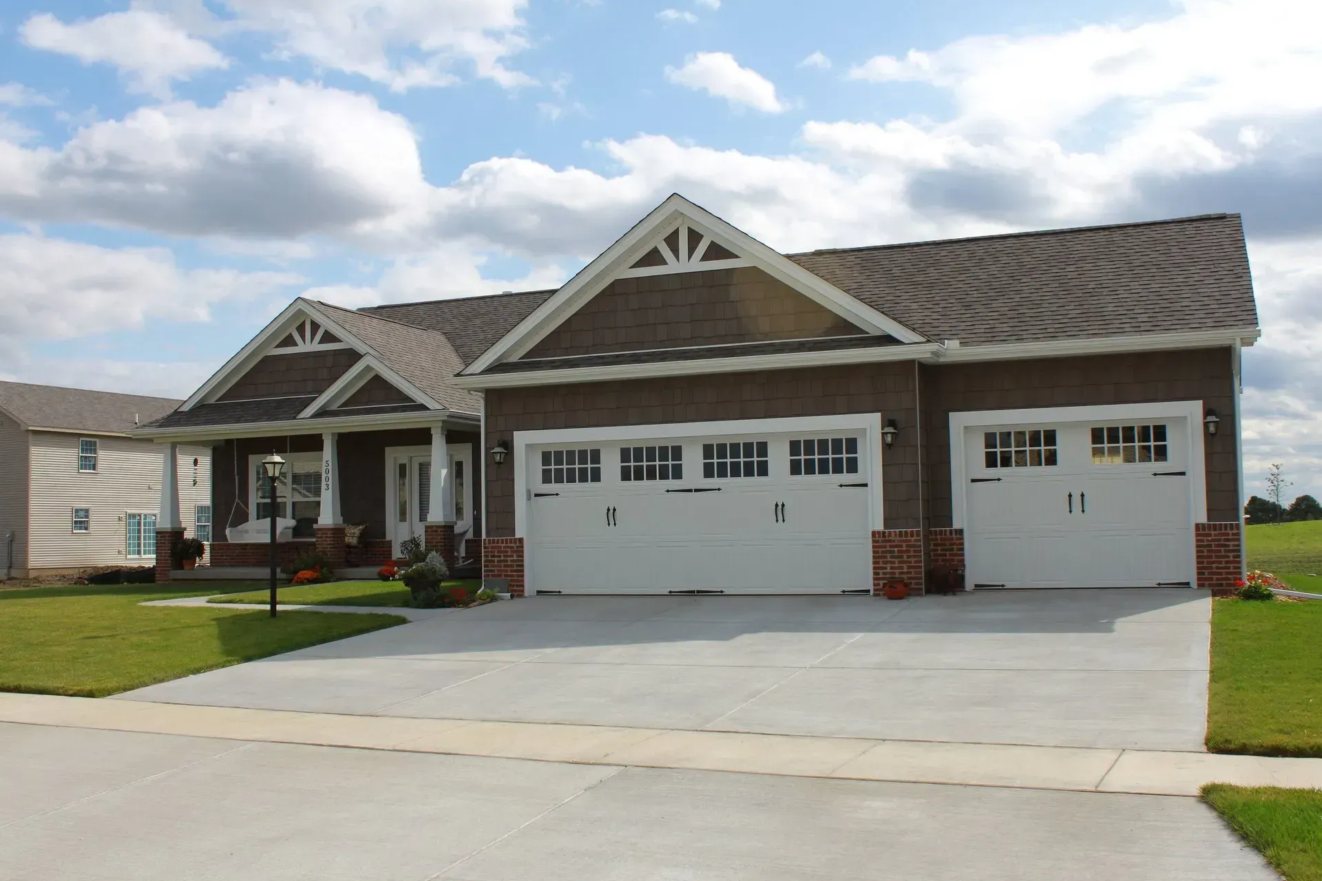 Brown house with white garage doors, driveway, and green lawn under cloudy sky.