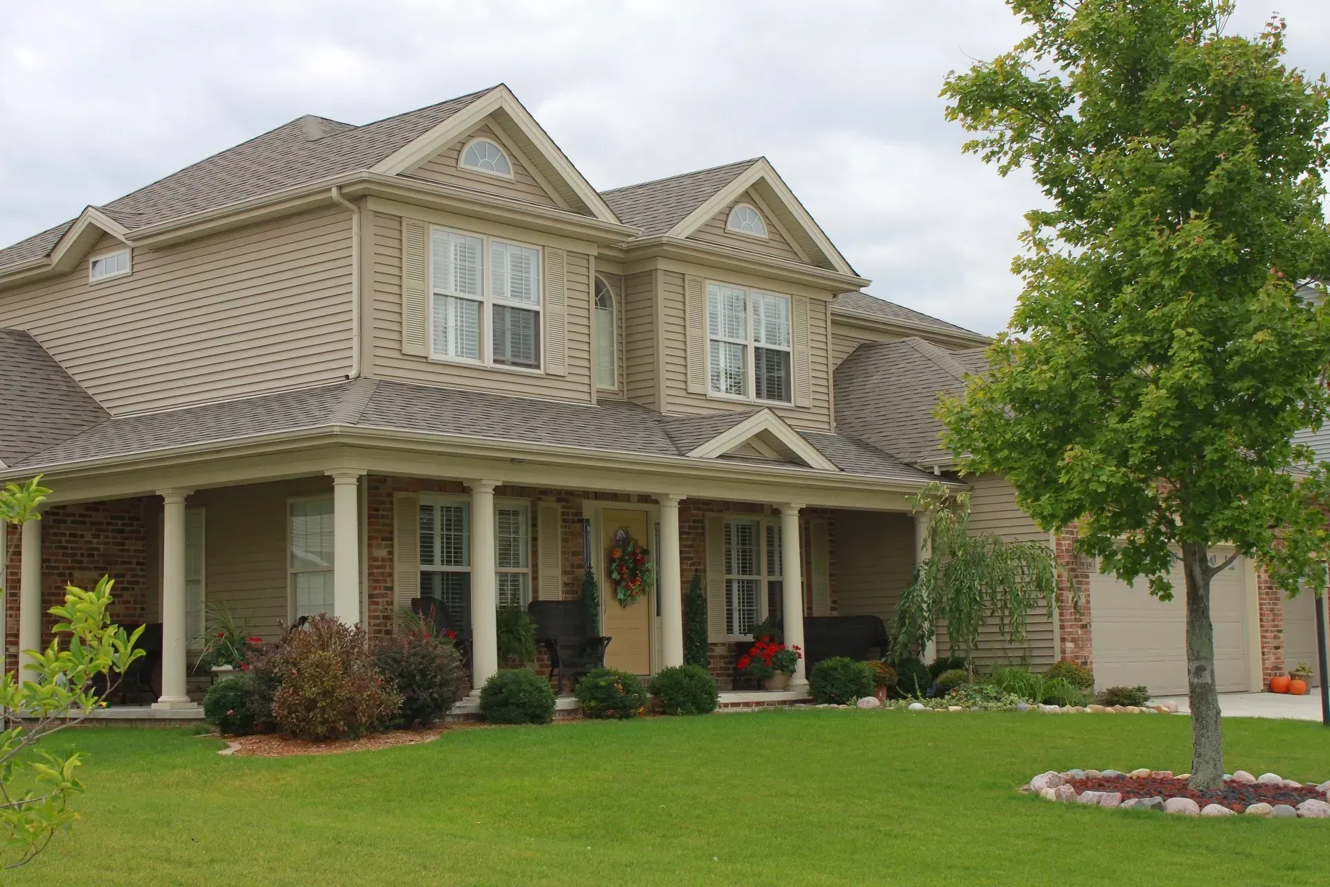 Two-story beige brick house with a covered porch, green lawn, and tree.