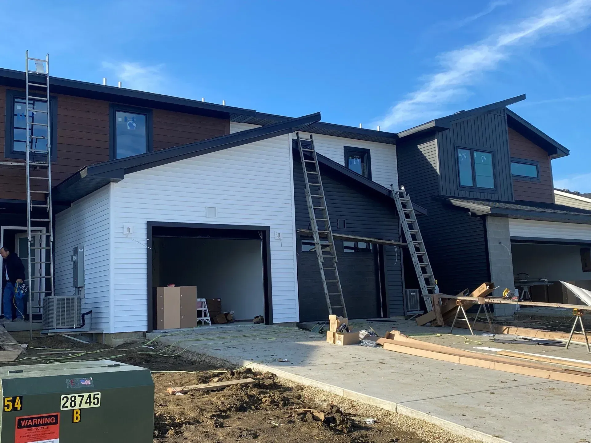 Construction of modern townhouses; white and black siding, garage, ladders, lift. Clear blue sky.
