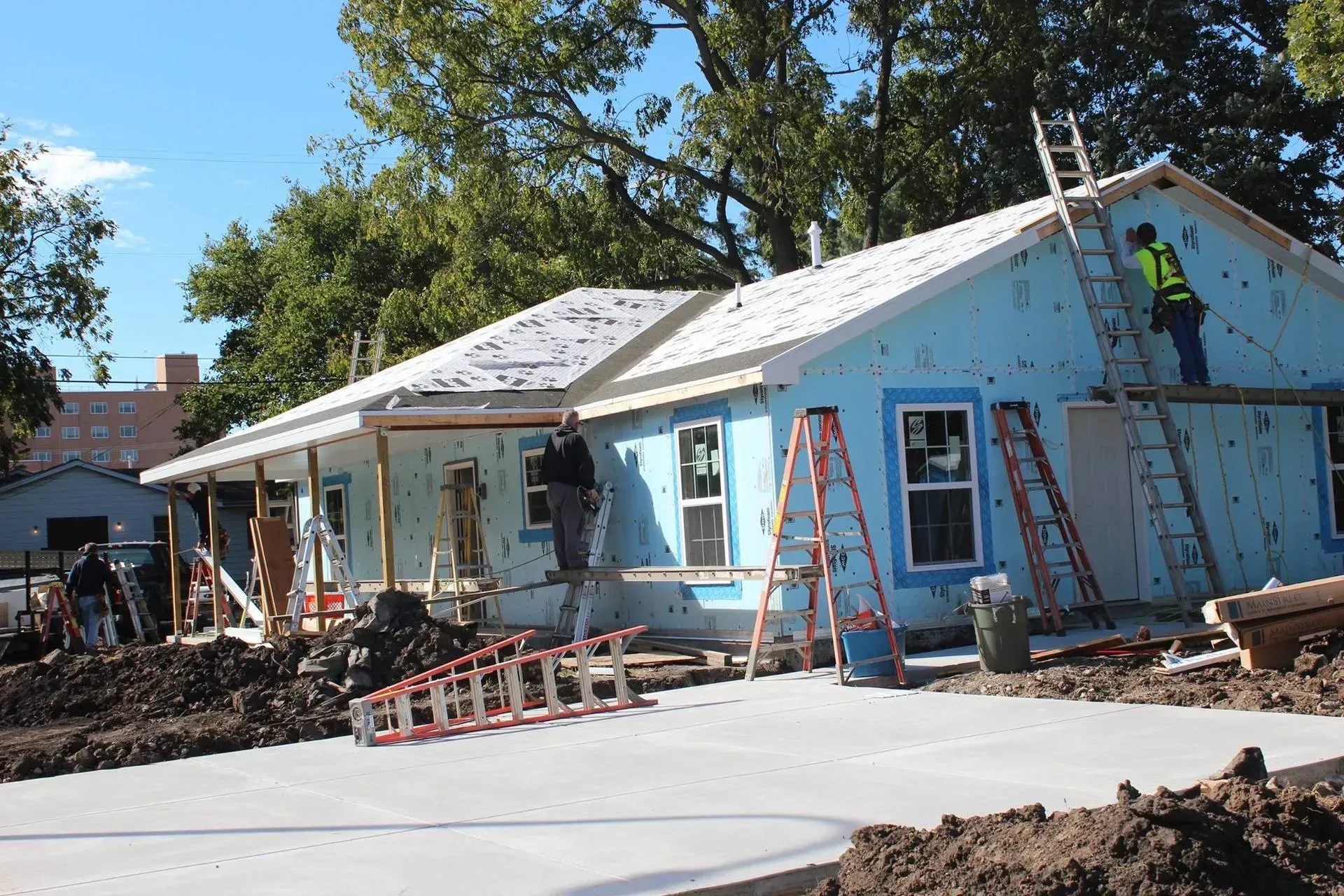 House under construction with workers on ladders, blue siding, concrete driveway, and clear sky.