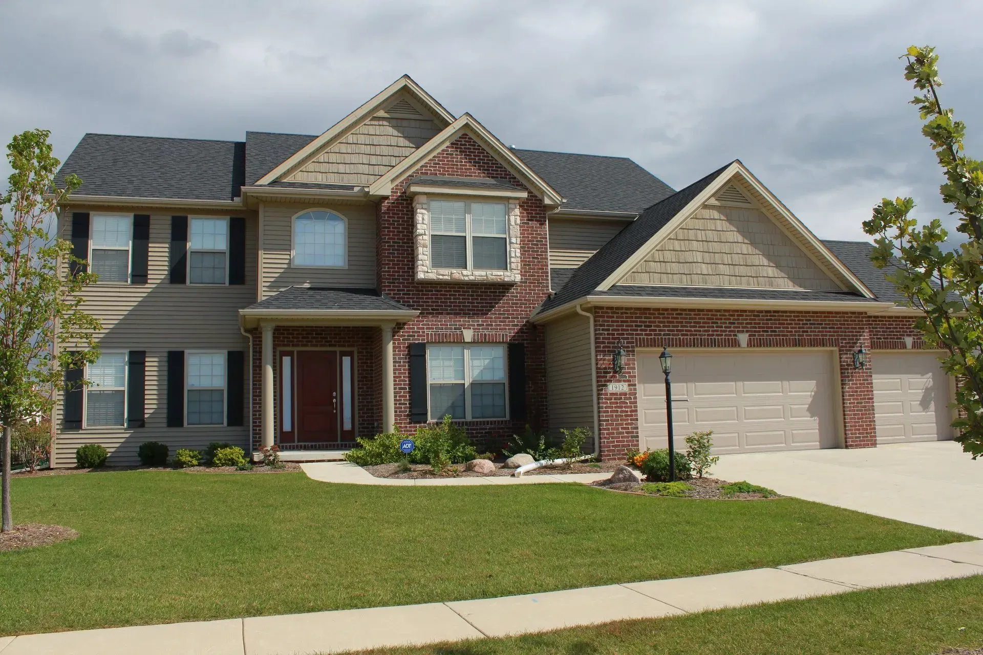 Two-story house with brick facade, beige siding, black shutters, three-car garage, and green lawn under a cloudy sky.