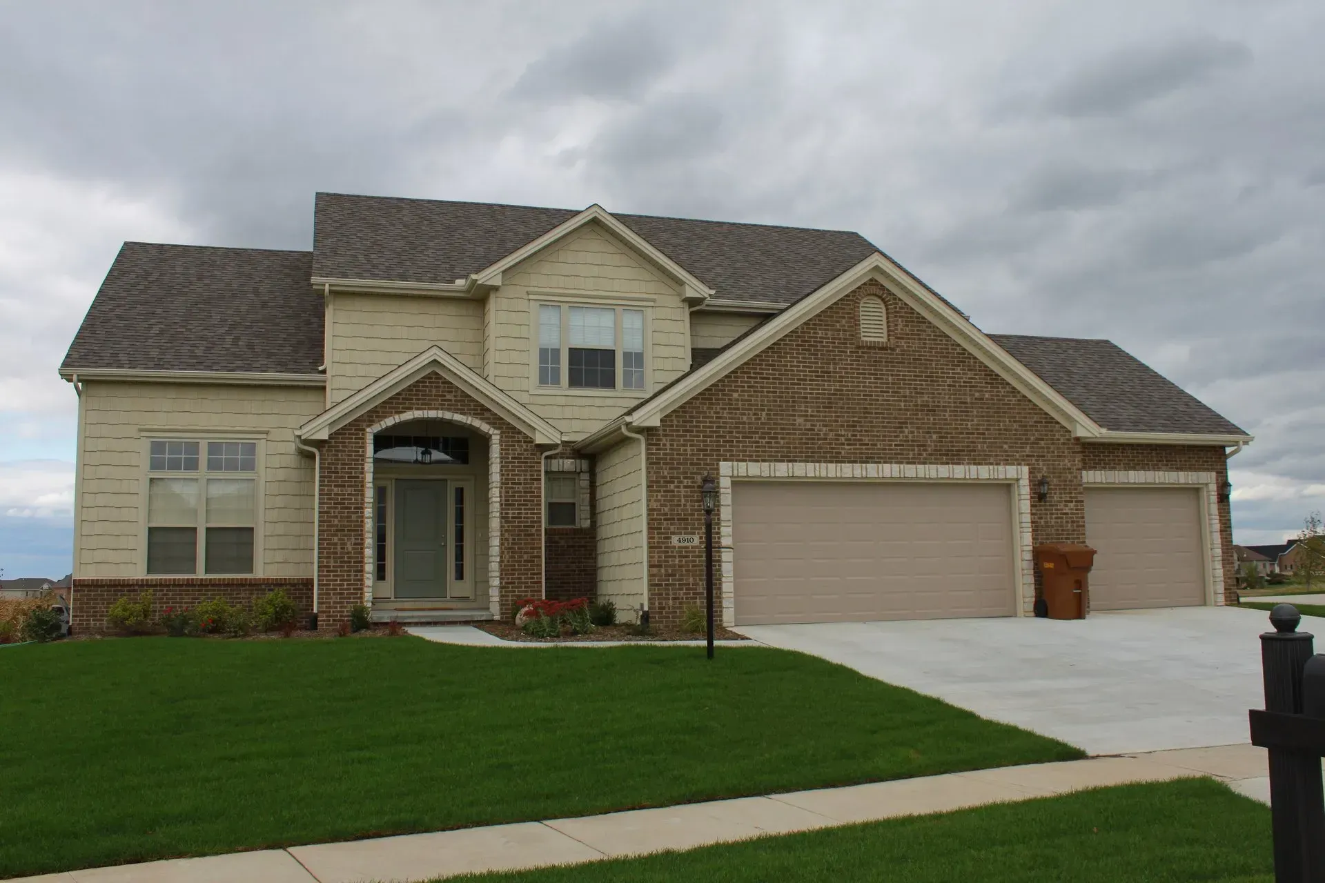Two-story beige and brick house with a green lawn, two-car garage, and cloudy sky.