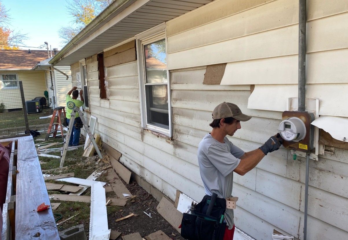Two workers in work clothes perform maintenance on the exterior of a light-colored house, one removing an electric meter.
