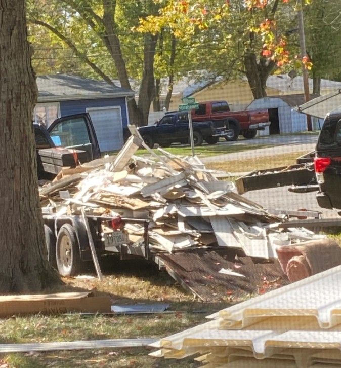 A trailer parked on a lawn is piled high with construction debris, including wood and white siding, near a tree and road.