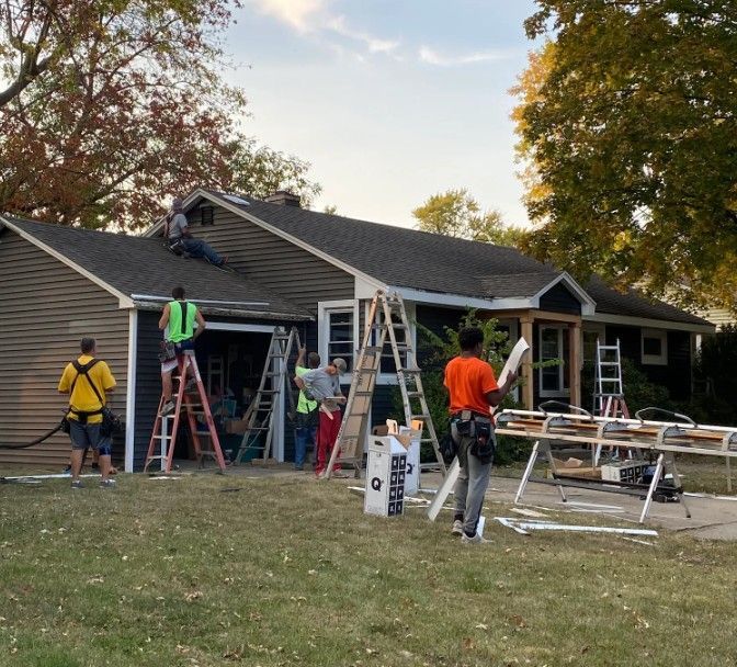 A crew works on a home exterior, installing siding and roofing from ladders on a sunny day.