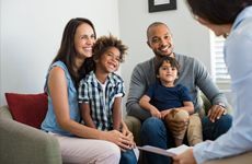 A smiling family of four sits on a living room couch, engaging with a person across from them. The scene conveys warmth and happiness.