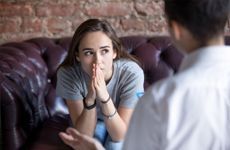 A woman sits on a burgundy sofa, looking pensive with hands clasped near her face. She listens to another person, suggesting a serious conversation.