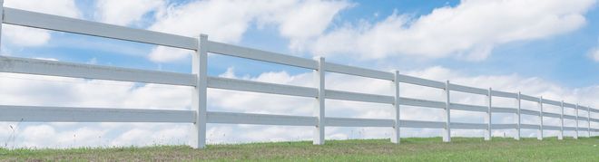 White wooden fence stretching across a grassy field under a partly cloudy blue sky.