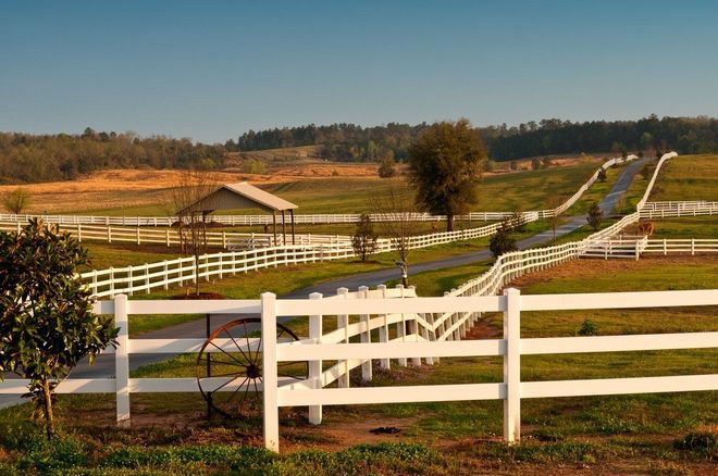 White fences border a rural dirt road leading through fields to a small barn under a clear sky