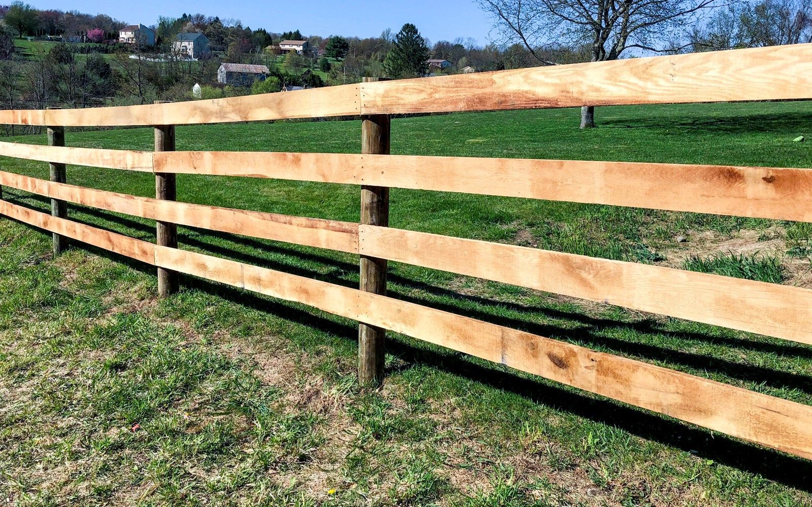 Wooden fence with three horizontal rails along a grassy field on a sunny day