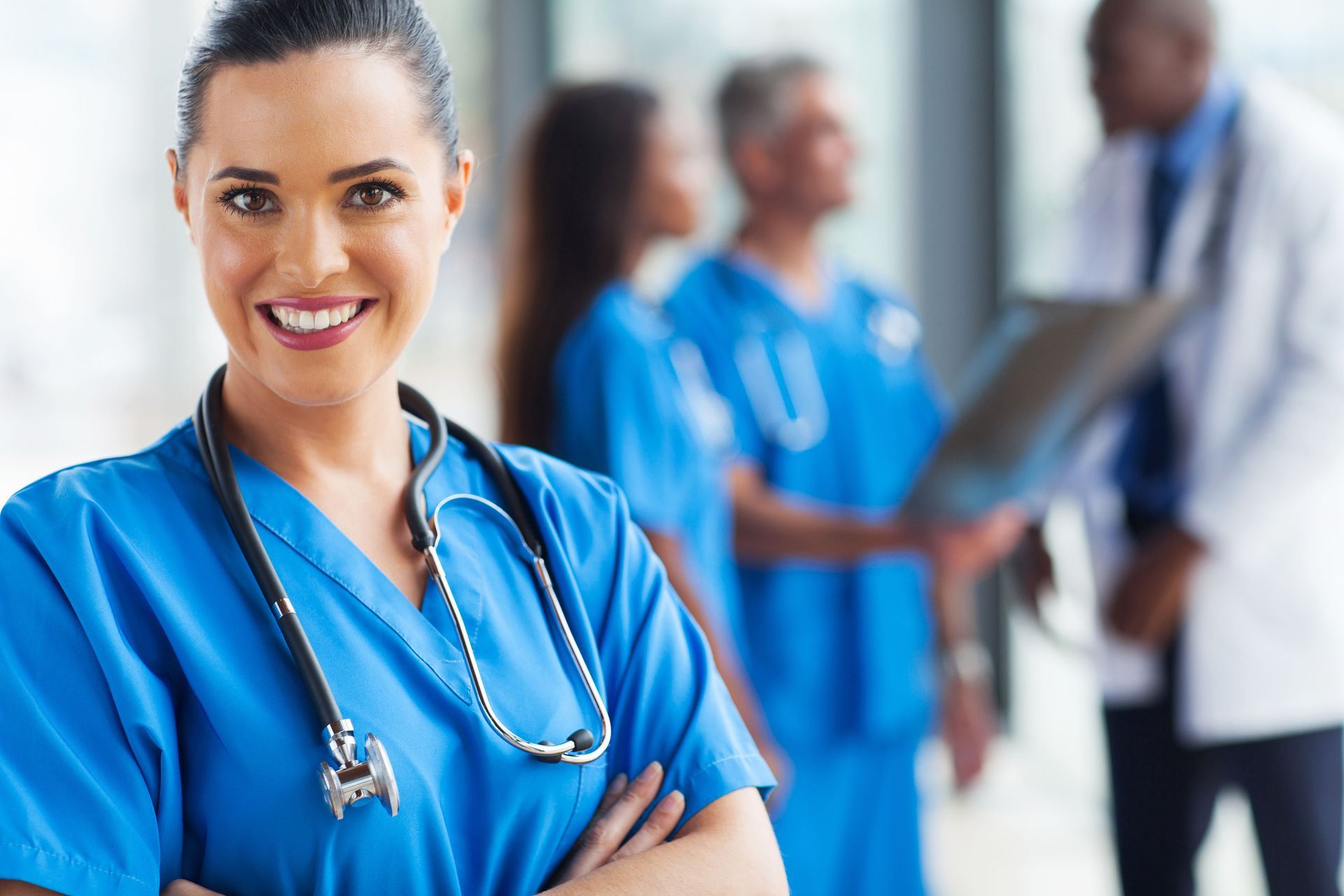 Smiling nurse in blue scrubs with stethoscope, standing in a hospital hallway with doctors blurred in the background