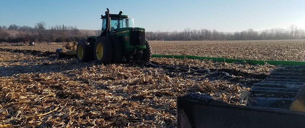 Green tractor plowing a harvested corn field under a clear, sunny sky.