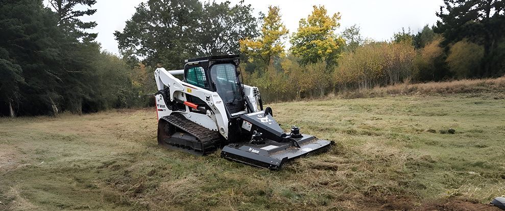 A Bobcat skid steer with a mower attachment is parked in a grassy field, surrounded by trees.