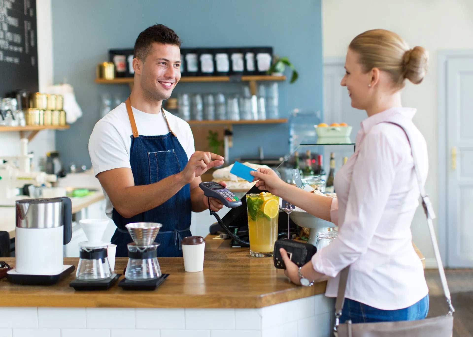 A barista in a blue apron smiles at a customer as she pays with a credit card at a cafe counter.