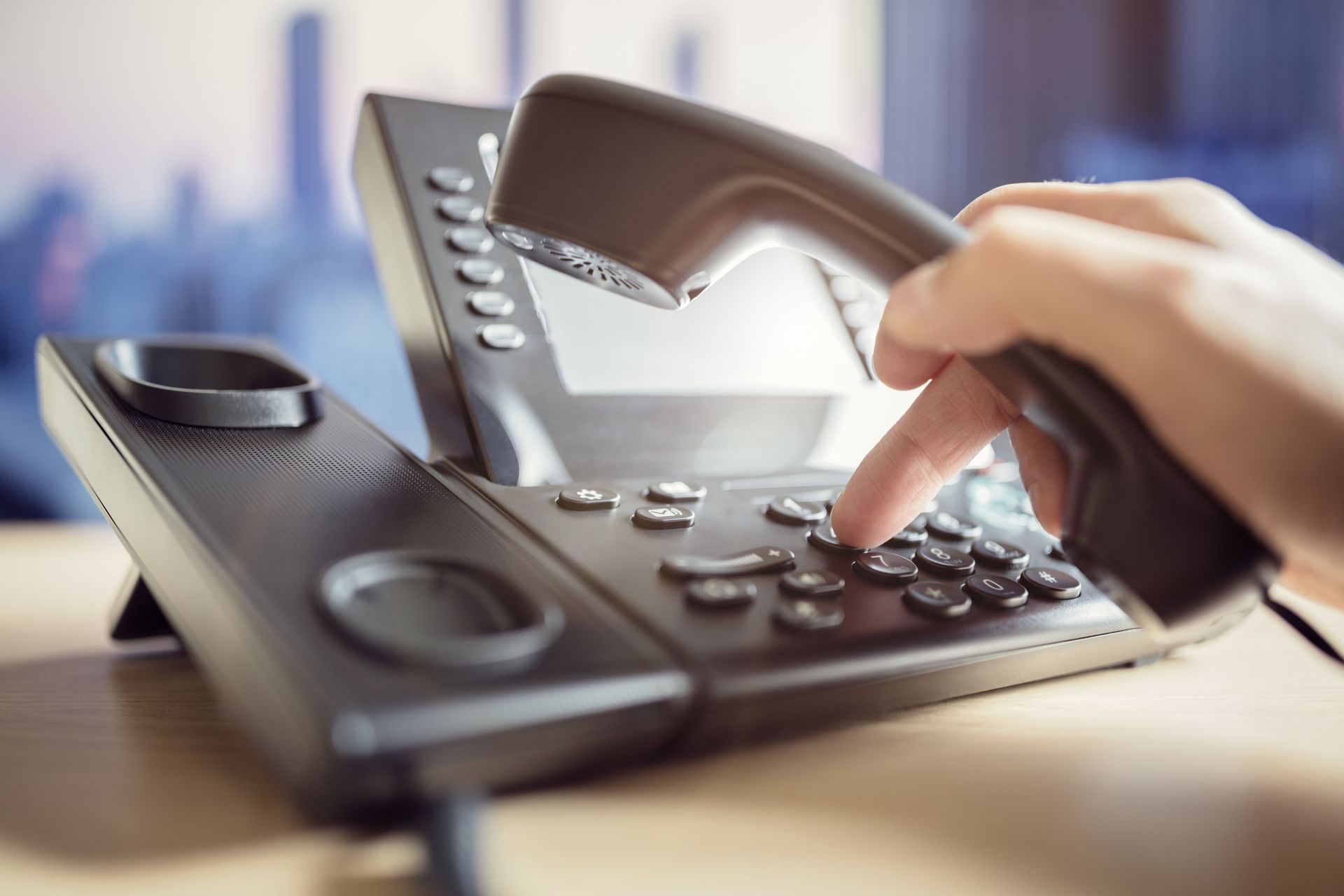A person's hand presses a button on a black office telephone while holding the handset to their ear.