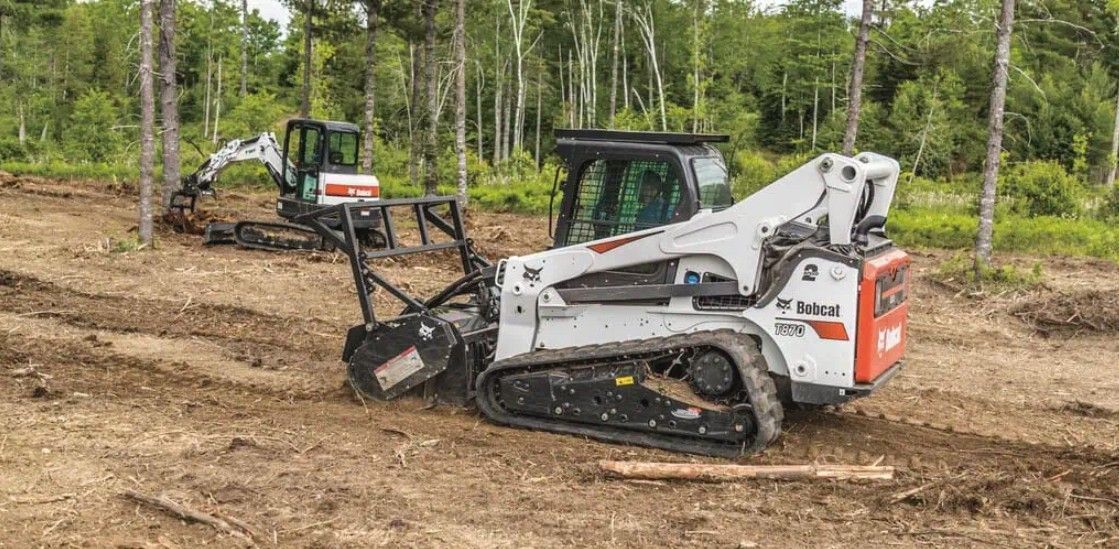 Two compact excavators clearing dirt in a wooded construction site.