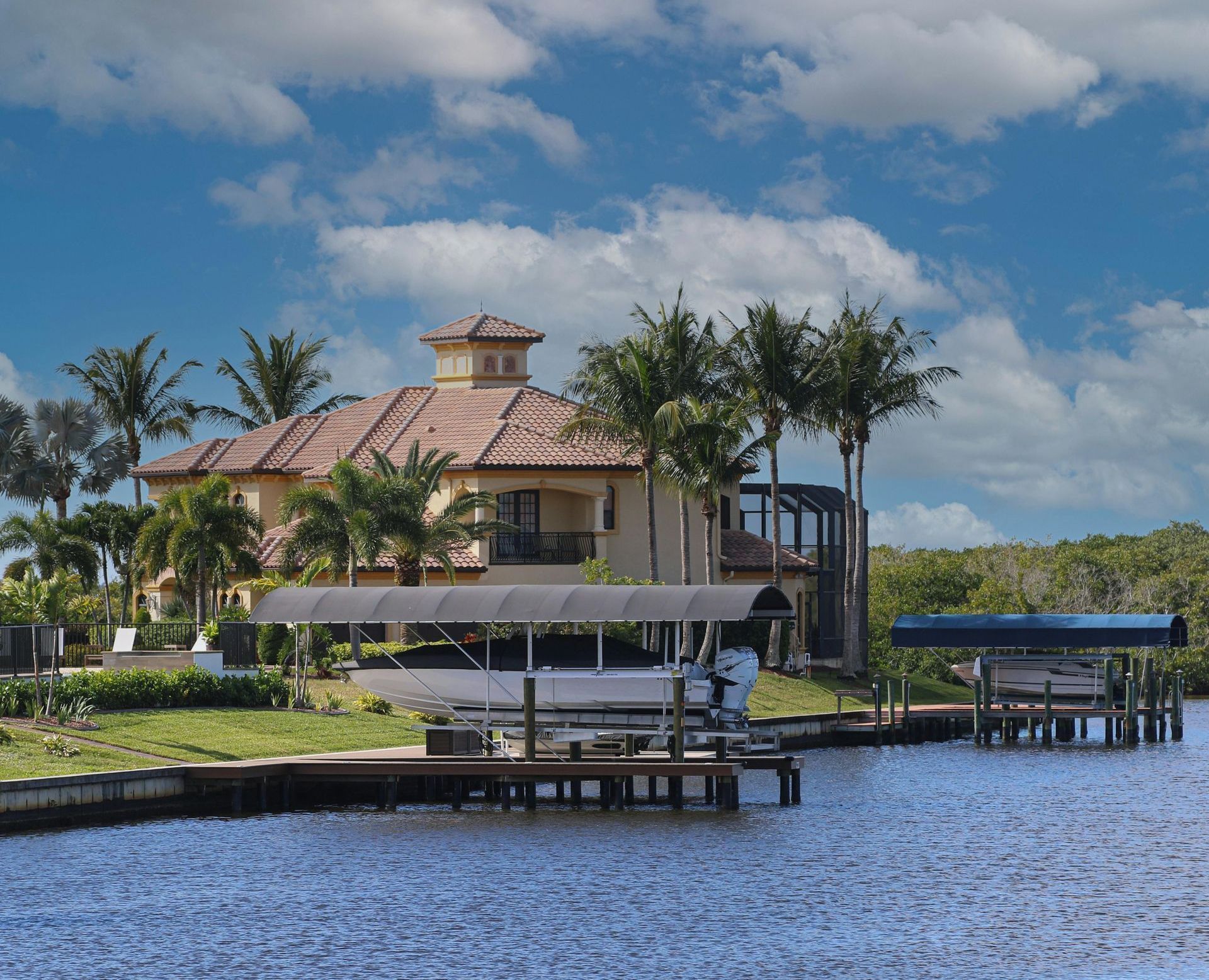 Luxury waterfront home with boat docks, blue water, and palm trees under a cloudy sky.