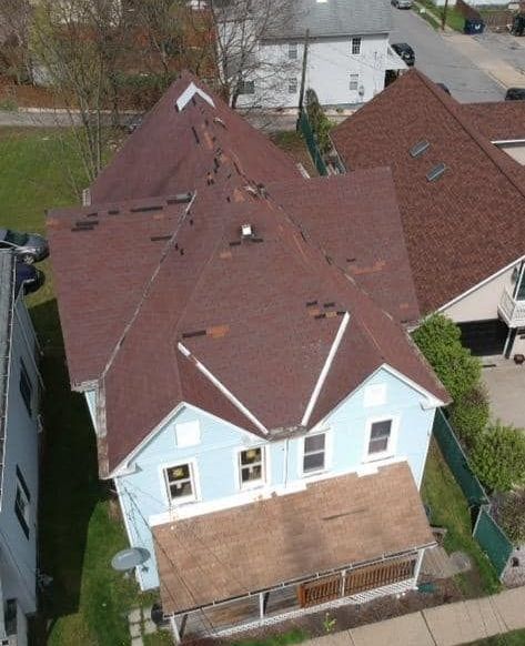 Aerial view of a blue house with brown roofing and a porch. Houses and cars are in the background.